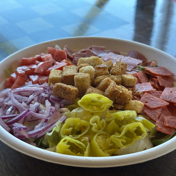 A large salad with tomatoes, red onions, croutons, pepperoni, ham, and banana peppers.