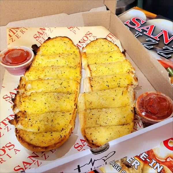 Santino's Pizzeria garlic breadsticks, golden brown with herbs, served in a box with two cups of marinara sauce.